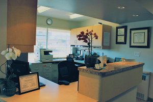 Modern dental office reception area featuring curved front desk, computer workstations, floral arrangements, and framed certificates on wall.