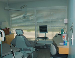 Dental treatment room equipped with blue patient chair, overhead dental light, computer monitor, cabinetry, and window with blinds.